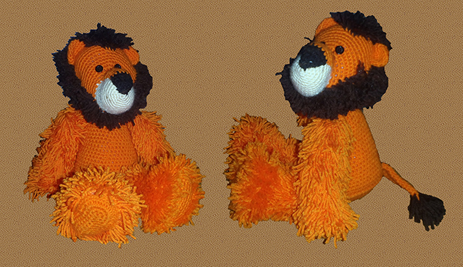 orange crocheted lion with a brown mane, white snout, black nose, a brown tipped tail, two button eyes, and furry yarn hair on all four legs.