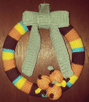 Fall/Thanksgiving Day Wreath using brown, orange, yellow, and olive colored yarns. 3 small crocheted pumpkins sit at the bottom of the wreath and an olive green crocheted bow is added to the top.