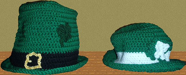 Green crocheted top hat with black band and yellow buckle adorned with crocheted clovers. Also a green crocheted trilby hat with white band and adorned with a green and white clover